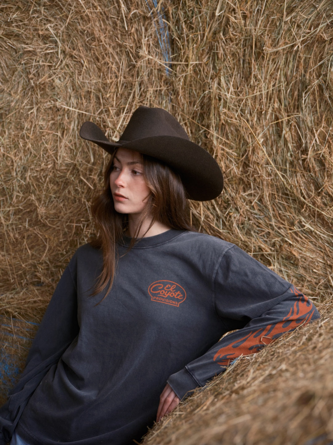 Woman wearing a faded black longsleeve t-shirt with a red script logo, leaning on a hay bale.